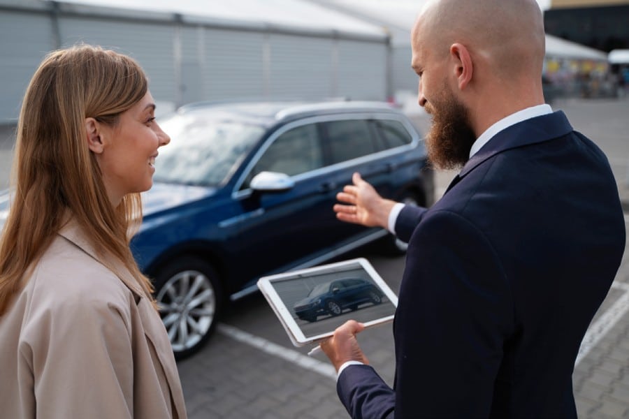 Woman engaging with a car dealer, discussing vehicle features while he holds a tablet displaying a car image, outdoor dealership setting.