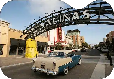 ```json { "alt": "A vintage car drives under the Salinas arch in a downtown area with shops and buildings.", "title": "Vintage car under the Salinas arch downtown" } ```