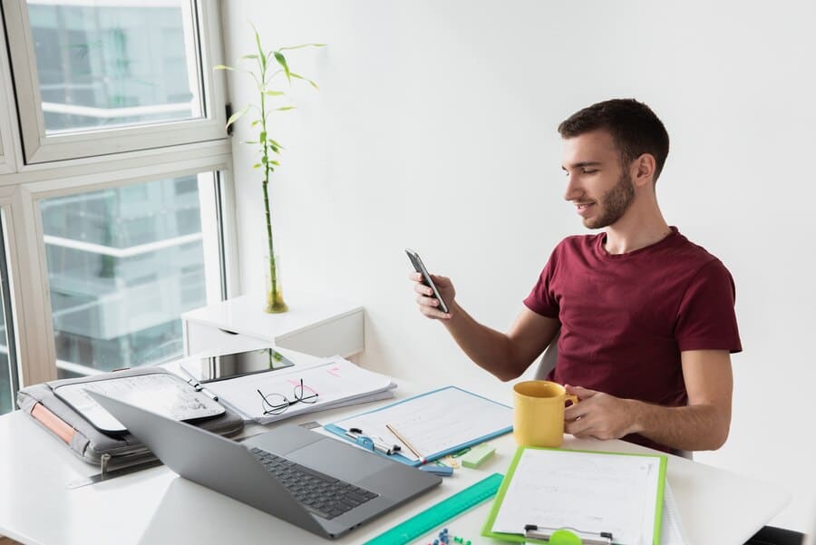 Man sitting at an office desk using a smartphone, with a laptop, documents, and a coffee mug, illustrating a Bring Your Own Device (BYOD) work environment.