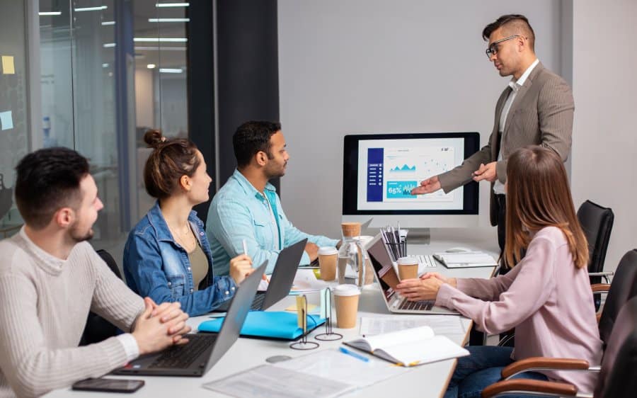Business meeting with diverse professionals discussing managed IT services, featuring a presenter pointing at a screen displaying performance graphs and statistics, laptops and coffee cups on the table.