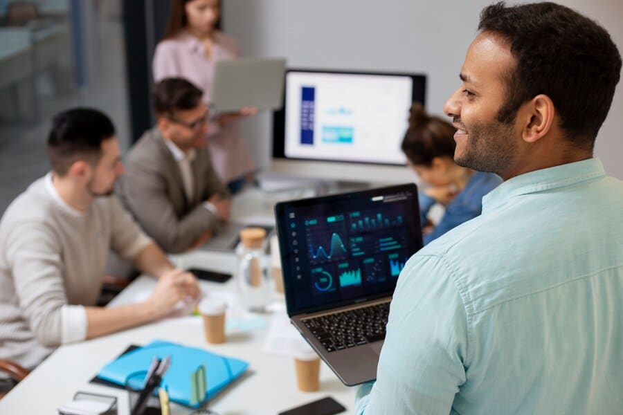 Business meeting with professionals discussing IT solutions, one man holding a laptop displaying data analytics, multiple screens in the background showing graphs and charts, collaborative workspace environment.